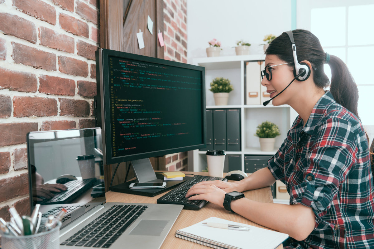 A women in her home office working on code on her computer while wearing a headset and smiling