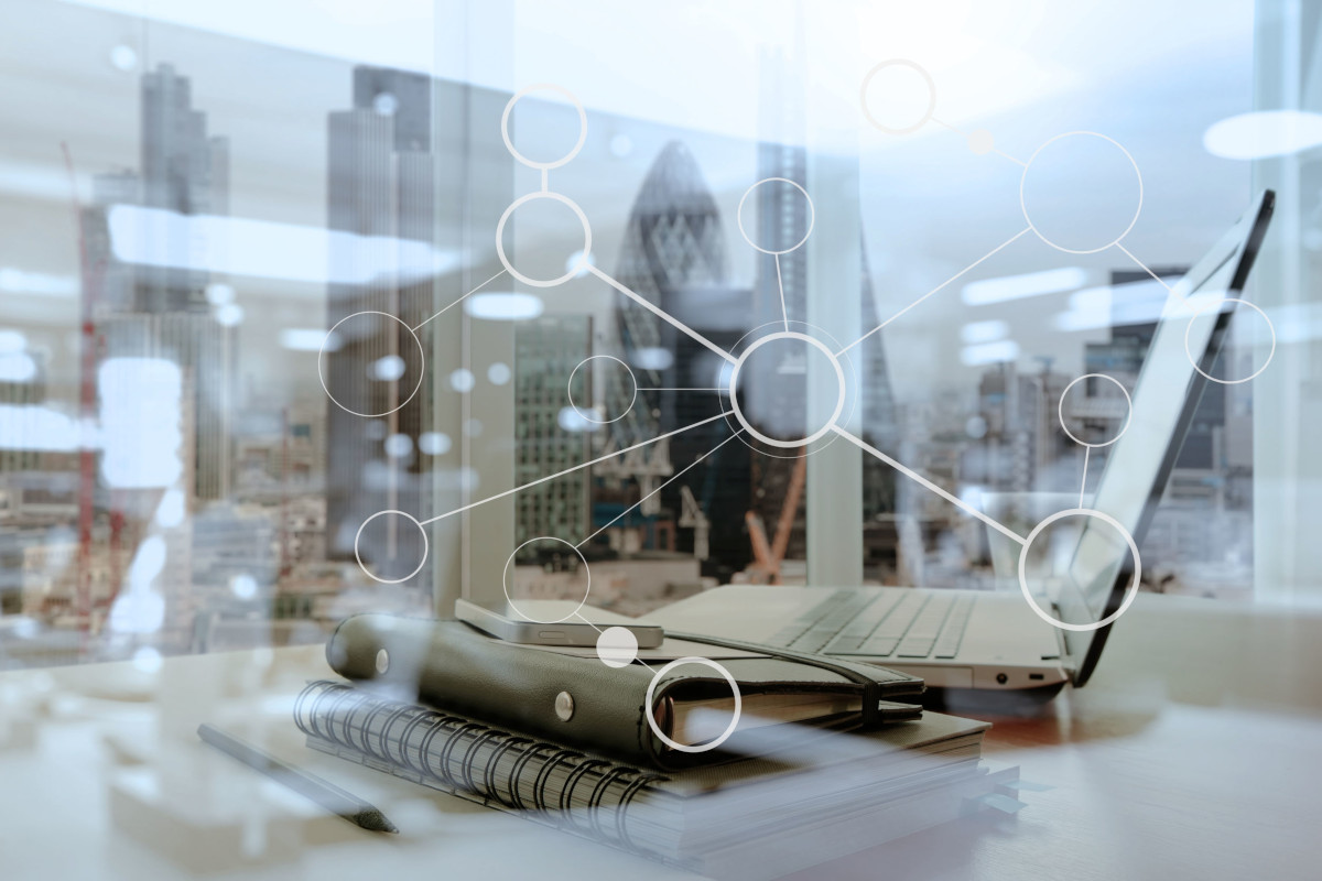  Close up of a laptop on a desk next to a stack of notebooks, with a city skyline and graphic of circles connected by lines imposed over it.