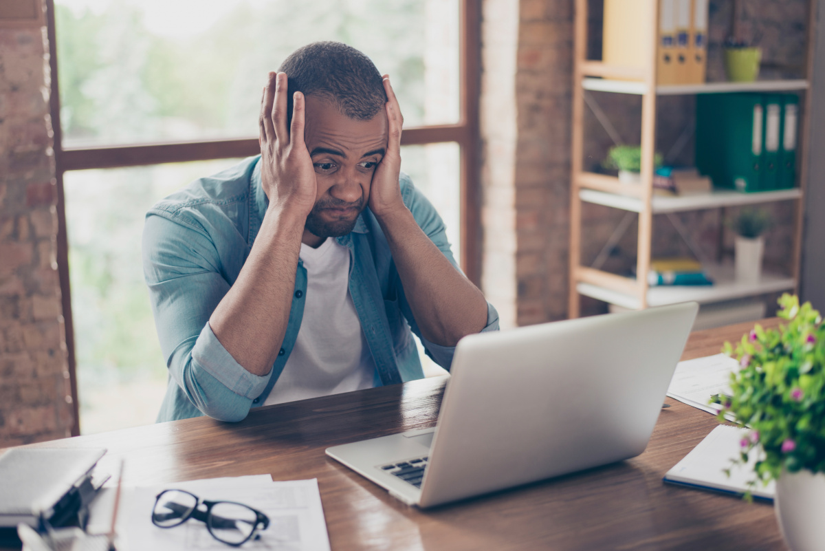 Frustrated worker holding face in hands while seated in front of laptop computer in office.